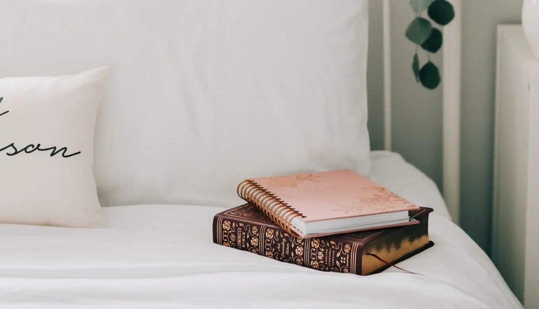 A journal and a Bible on white bed sheets next to a white pillow