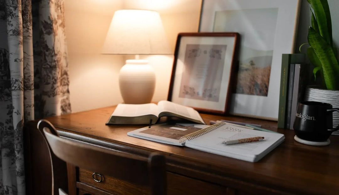 an open Daily Grace study next to an open bible and on a brown wooden desk, A lamp and some prints are on the desk against the wall