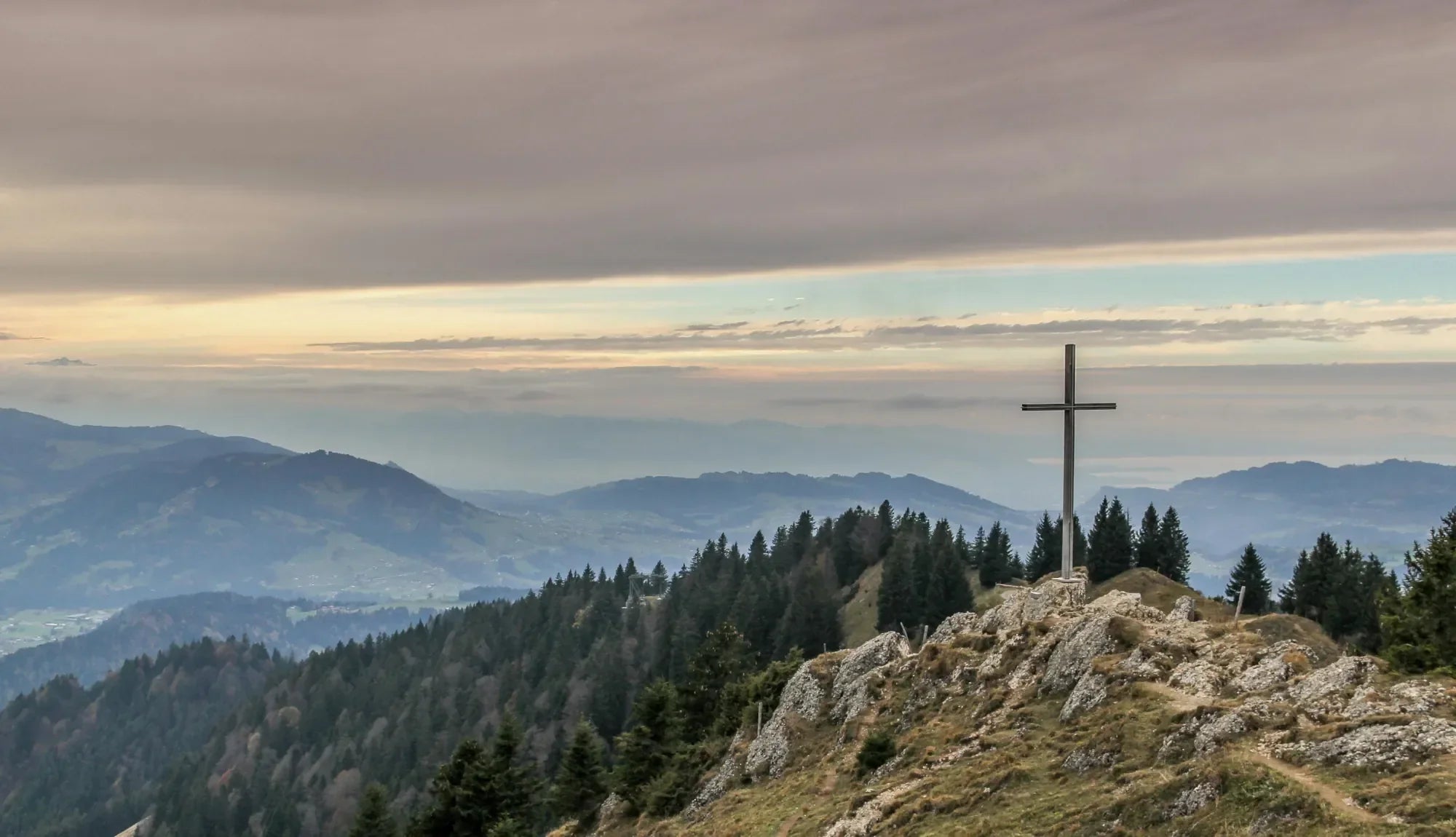 A Cross on a green and rocky hill overlooking a forest and mountains on a sunny day