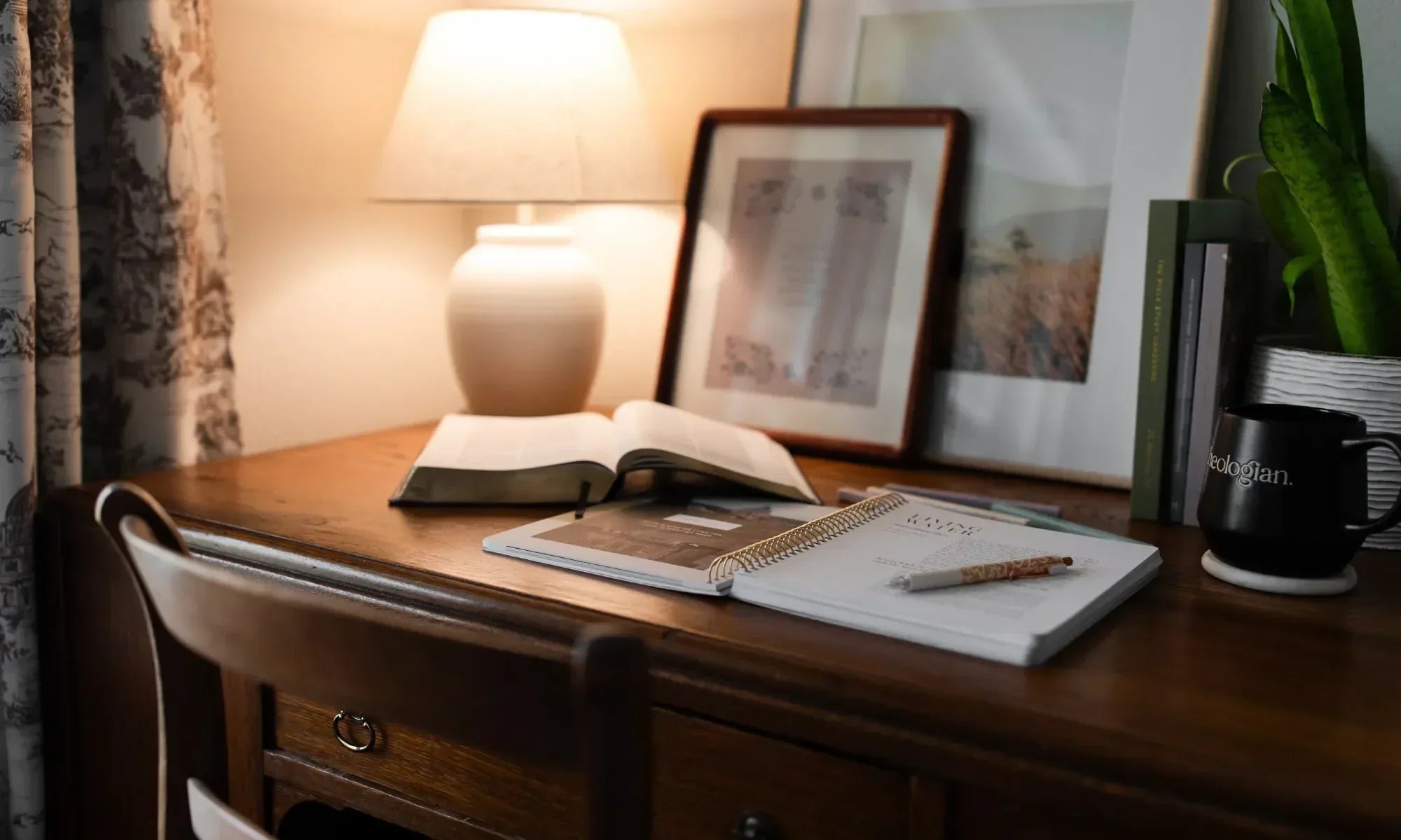 An open Daily Grace bible study near an open bible on a wooden desk with a lamp and some paintings near a white wall