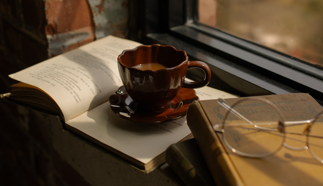 A brown coffee cup on an open book next to a pair of glasses resting on a book
