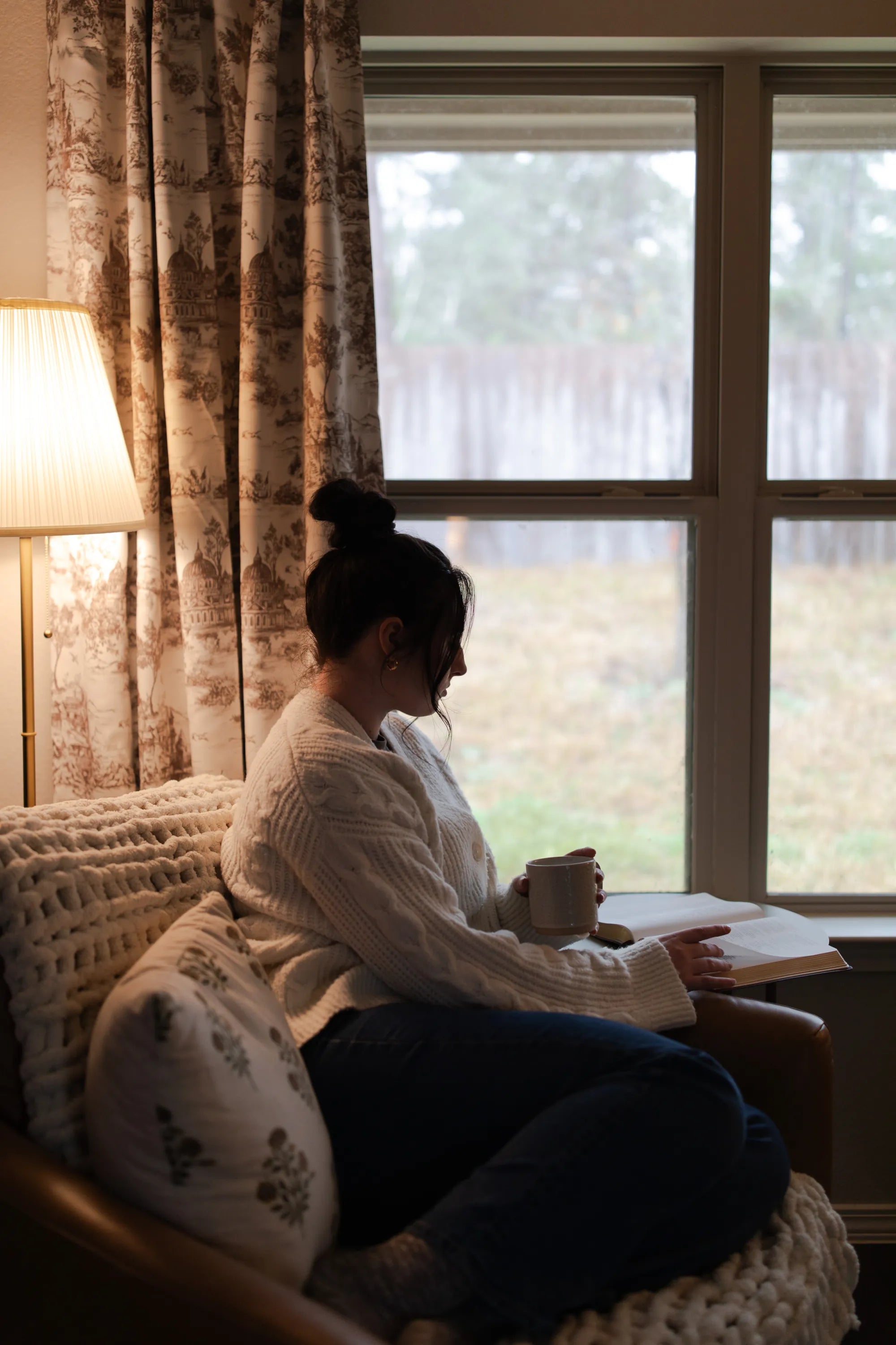 A woman reading her bible with a coffeecup in her hand next to a lamp and a window