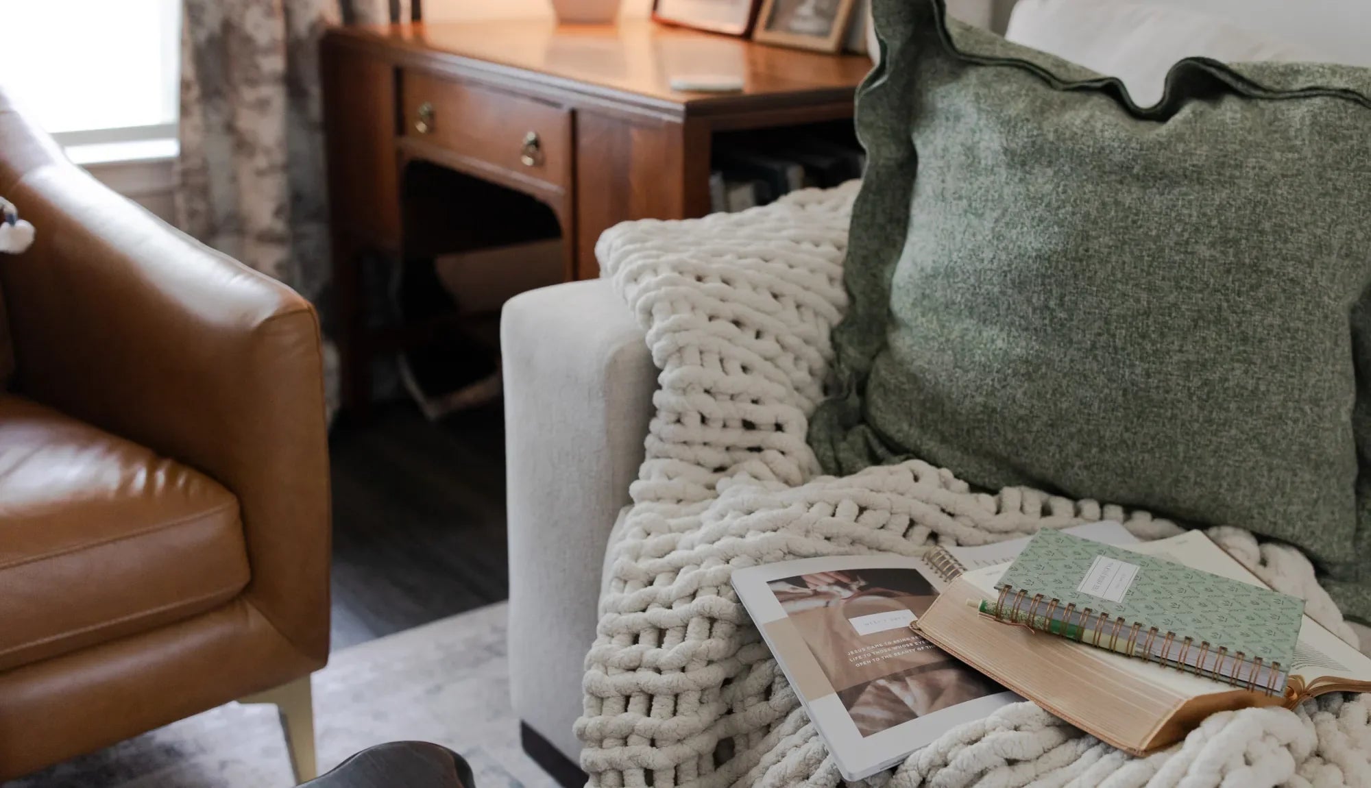 Multiple Daily Grace Products on a white couch on a white knitted blanket and a green pillow. Next to the couch is a wooden desk and a brown leather chair