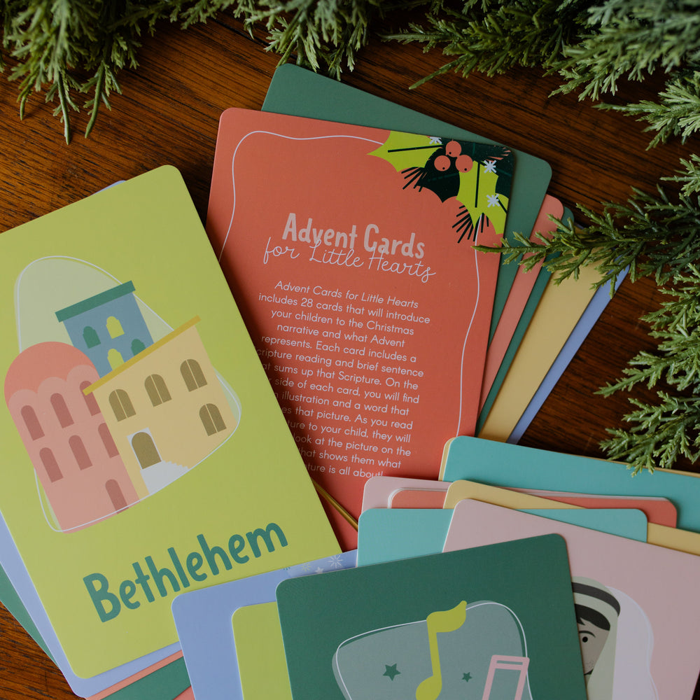 Multiple Advent Cards for Little Hearts stacked on a wooden table with greenery around it