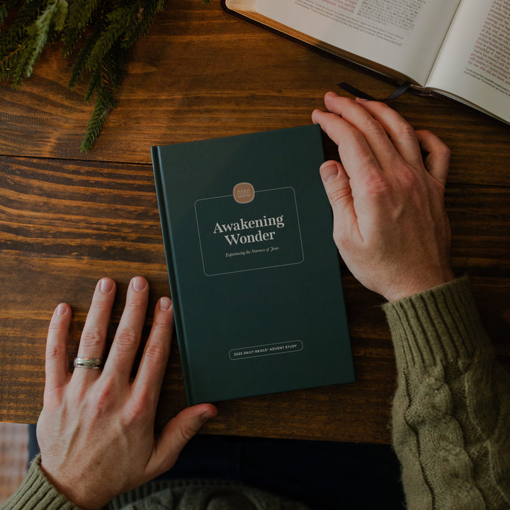 A man's hand holding the Awakening Wonder cover on a wooden table next to some greenery