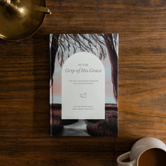 In the Grips of His Grace Study Cover on a brown wooden table next to a bronze oil lamp and a white coffee cup - Image 1 of 10