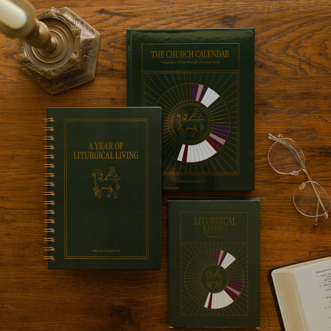 The Church Calendar, Year of Liturgical Living, and the Liturgical Living Cards laying on a wooden table next to a pair of glasses and a bronze candlestick