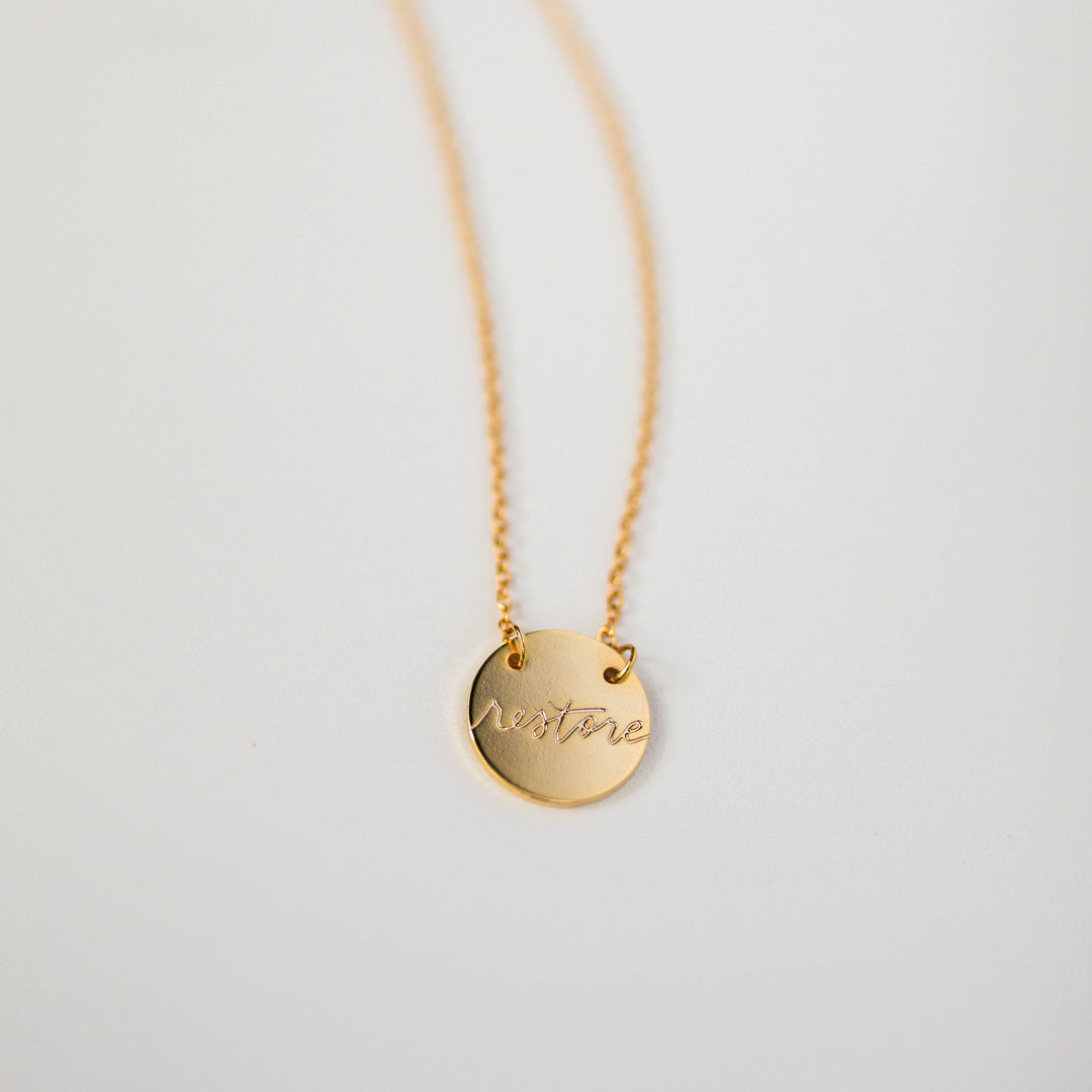 Gold necklace with the word "restore" written on circular pendant.
