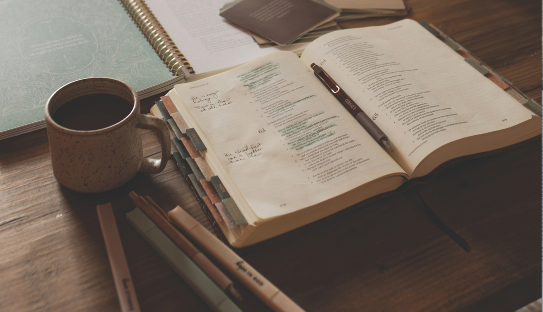 An open bible near multiple writing utensils and a coffee cup on a wooden table