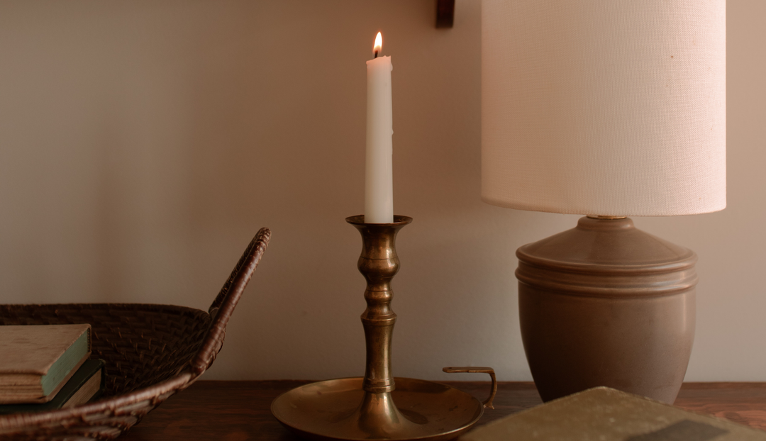 A single lit candle on a oil stand near a lamp and a small woven basket