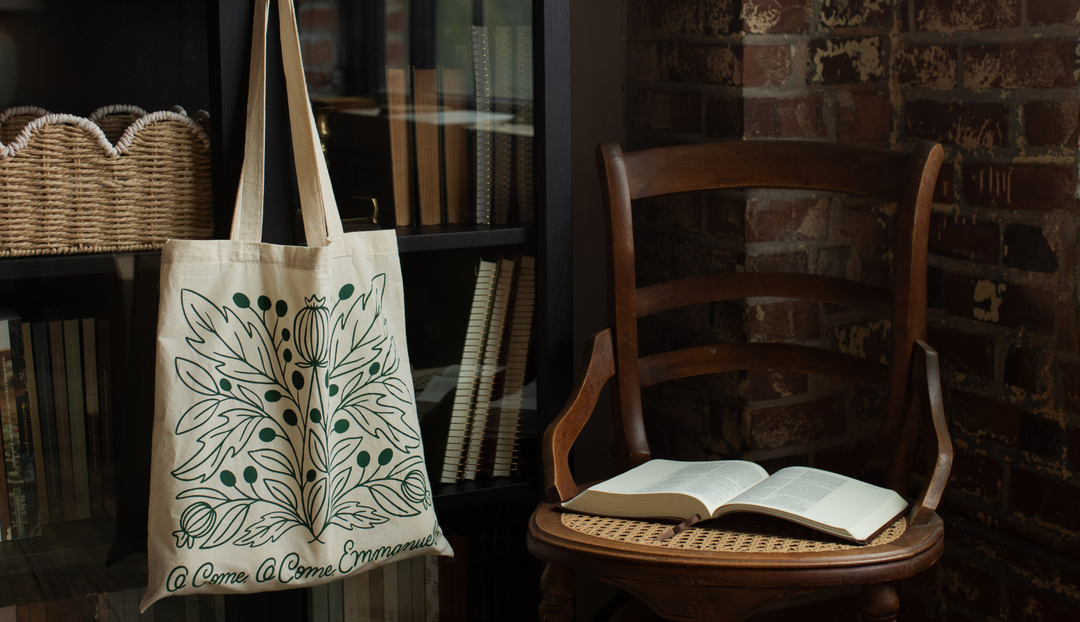 O Come O Come Emmanuel tote hanging on a black shelf next to a wooden chair with a open book on it