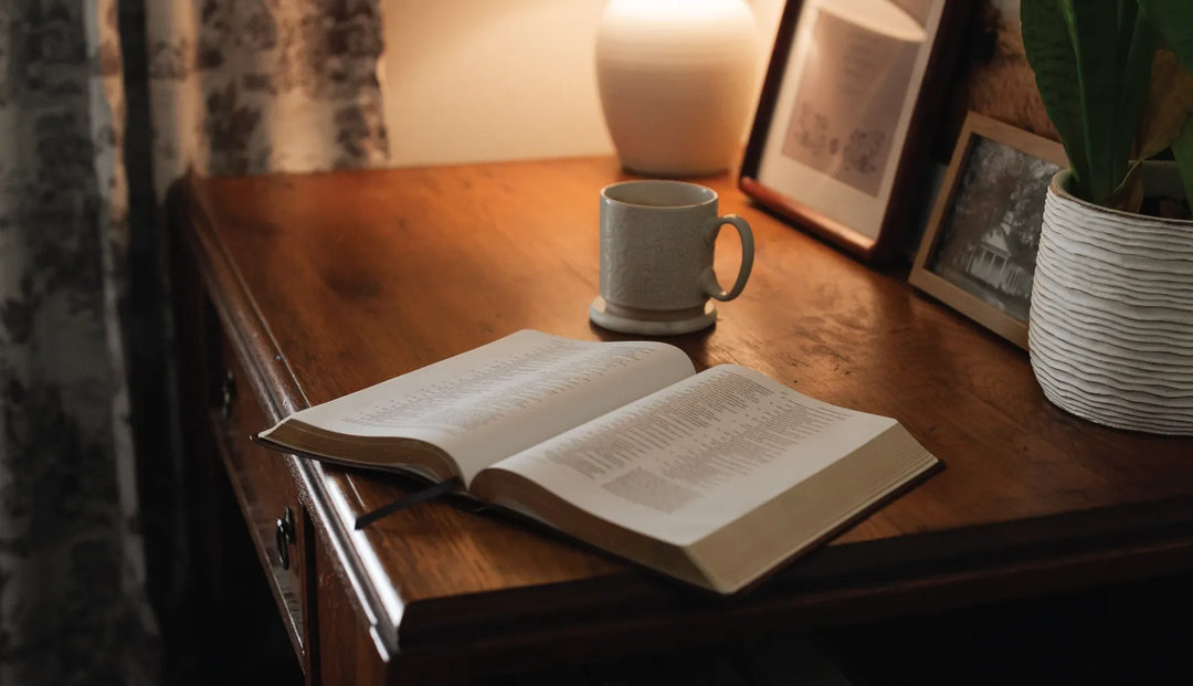 A cup of coffee next to an open bible on a side table next to some picture frames and a potted plant