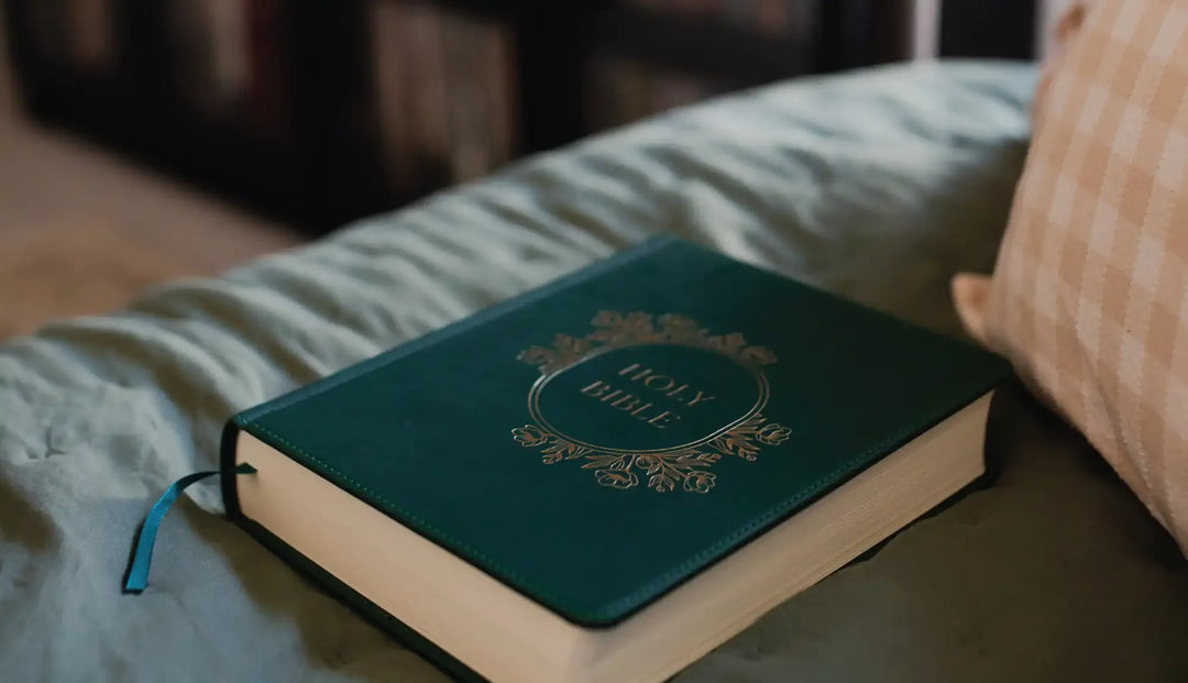 a green bible with gold etching laying on some blue sheets next to a yellow stripped pillow