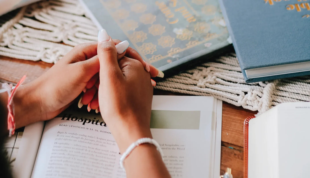 A woman holding her hands on top of a bible study with nearby bible studies on a knitted cover on a wooden table