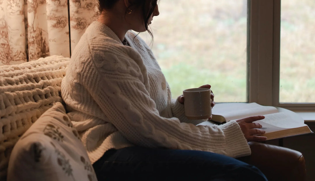 a woman wearing a white sweater holding a cup while reading a book 