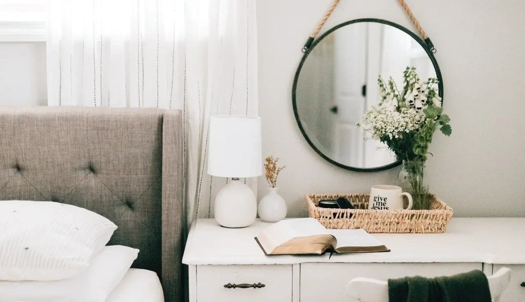 A bible on a desk next to a basket with a small white lamp nearby. A hanging mirror is next to white thin curtains behind a a bed with a gray frame headboard