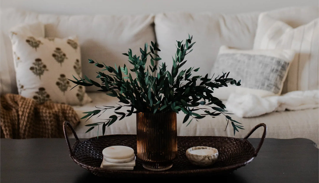 A potted plant on a stand on a black coffee table next to a light gray colored couch with two various styled pillows