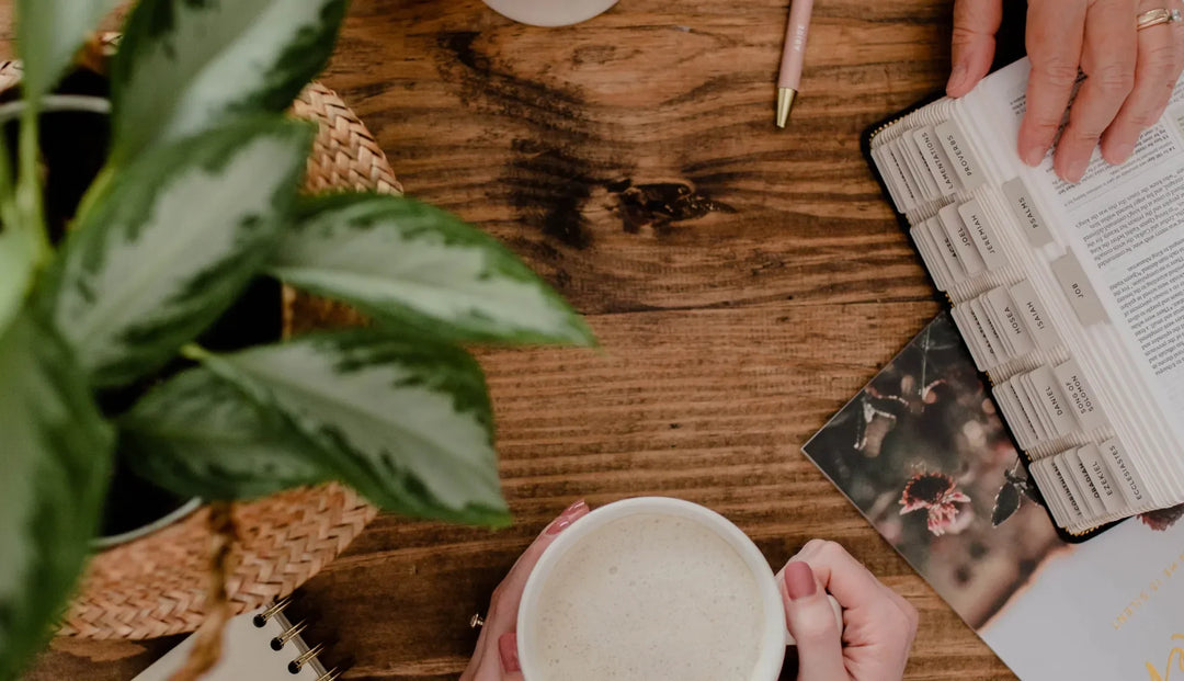 Two women's hands resting on a wooden table next to a bible and holding a coffee cup next to some greenery