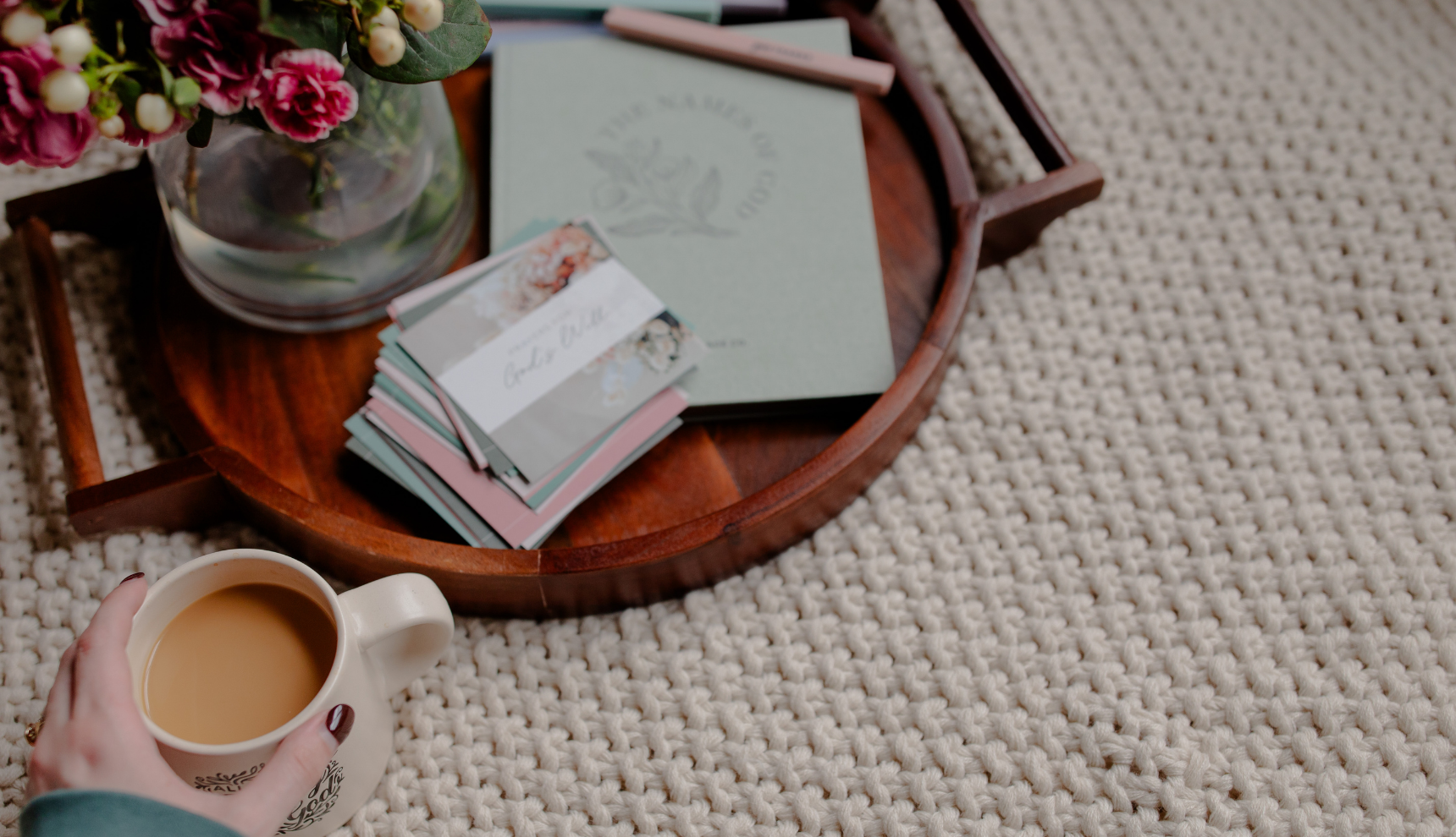 A woman's hand holding a coffee cup near a wooden tray with some verse cards and a vase of flowers on it