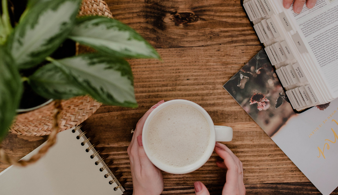 A woman's hand on a white coffee cup on a wooden desk next to a potted plant and various open books