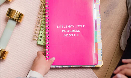 A pink journal labeled Little by Little Progress Adds up that is on a wooden desk with a woman's hand grabbing a corner of the journal