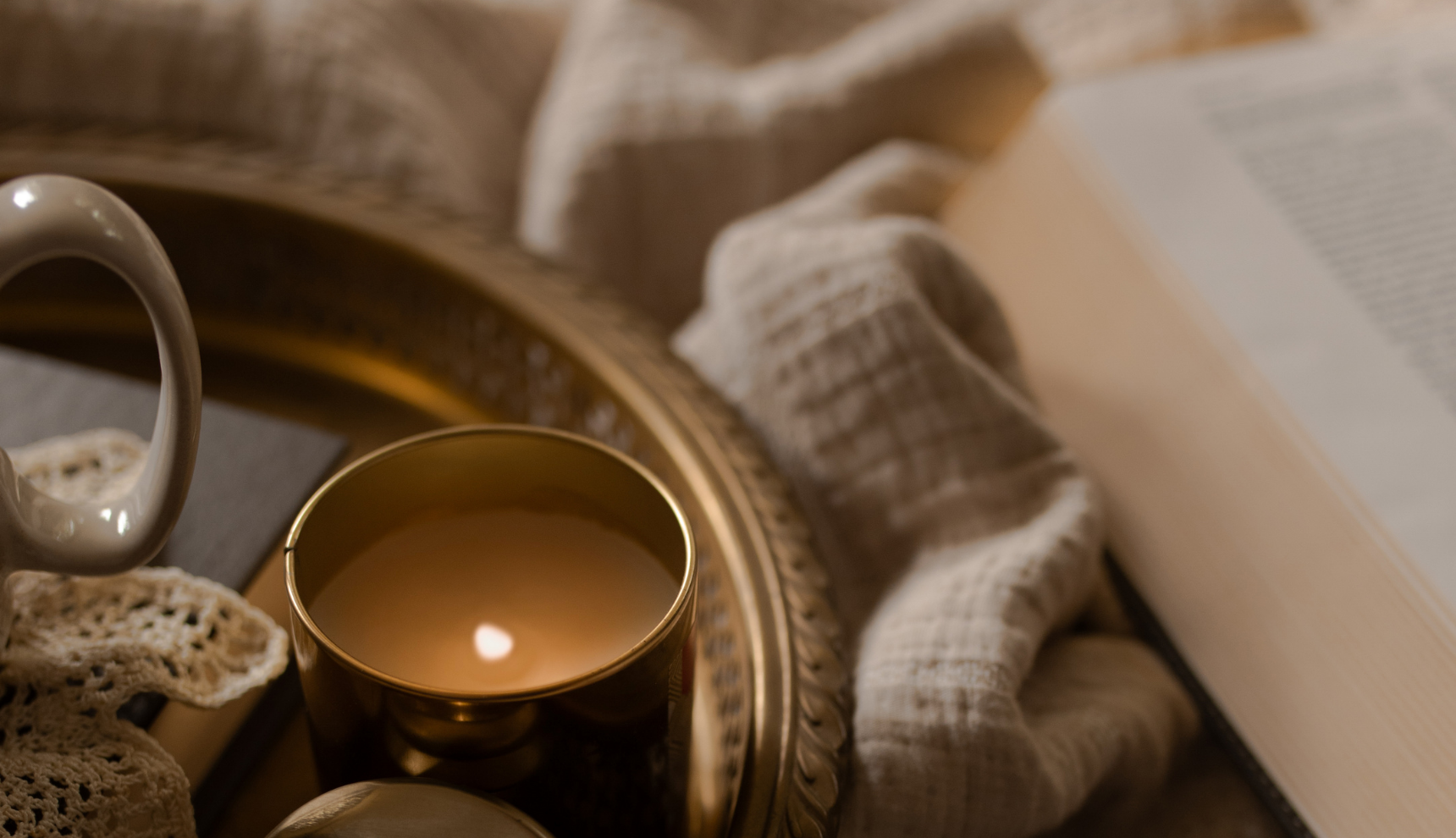 A lit candle on a tray next to an open book and a cream colored blanket