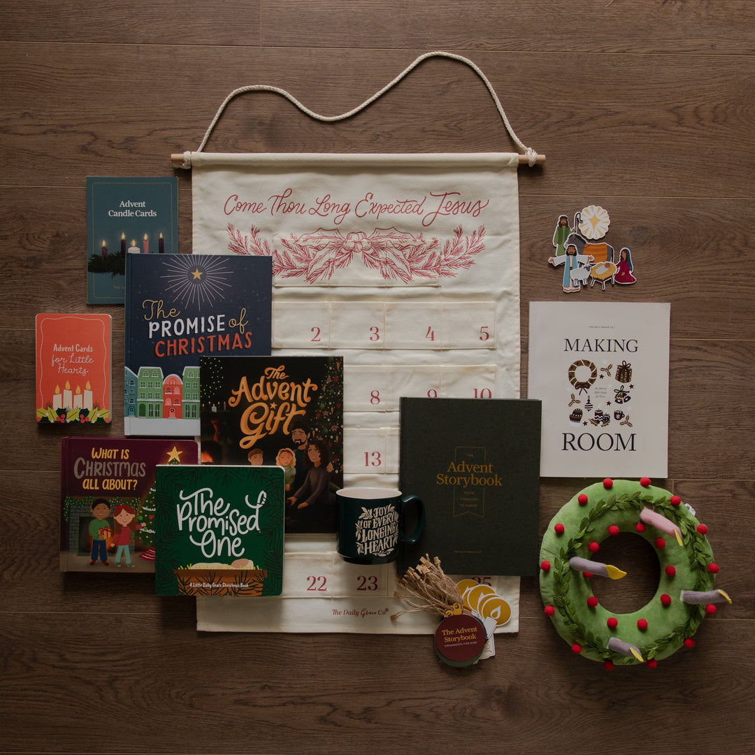 An Advent-themed gift bundle arranged on a dark wood background, featuring an off-white hanging calendar with red text and numbers, several Advent books and cards, a black mug, a green plush wreath with red ornaments, and other small holiday gift items.