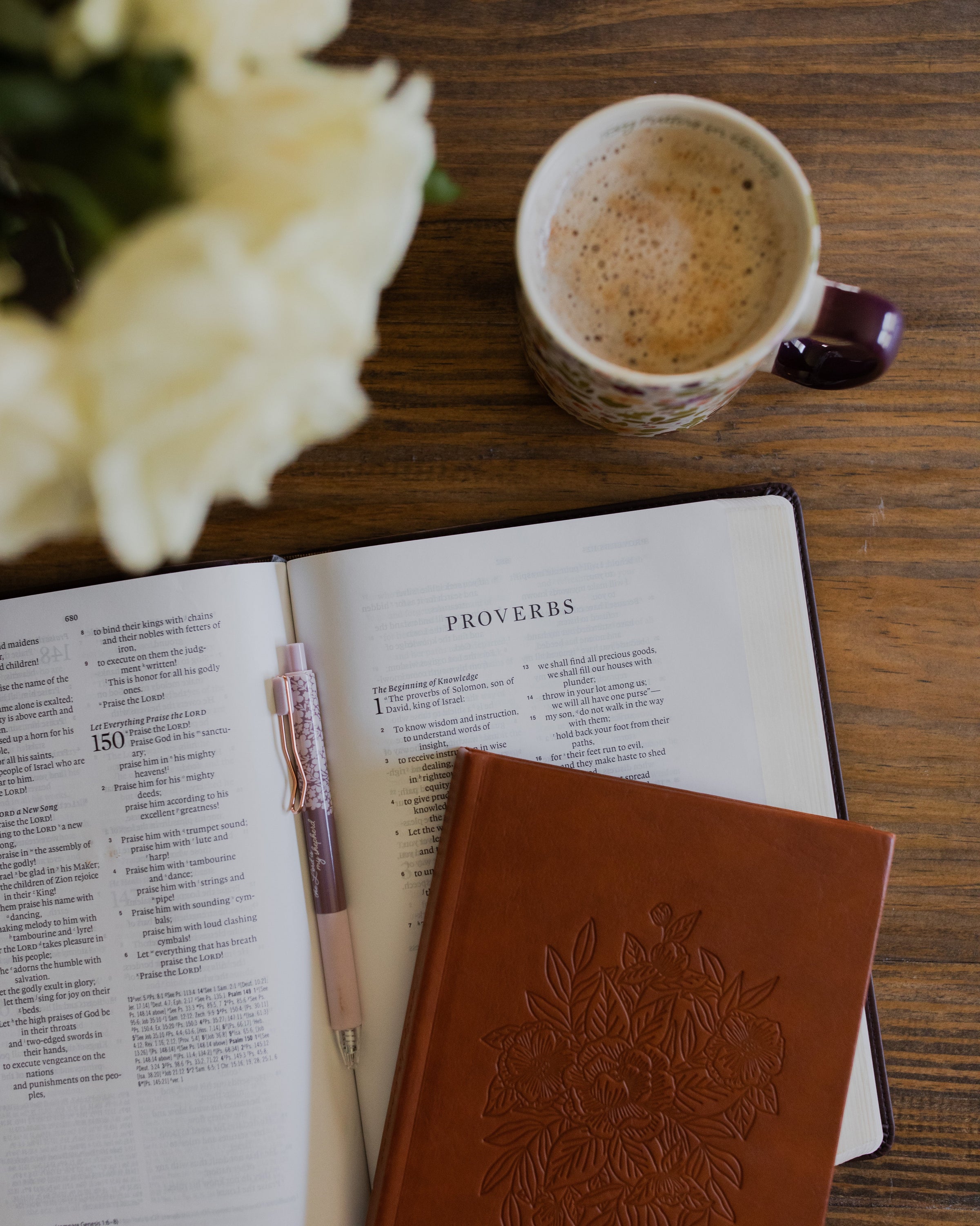 a closed leather journal on top of an open bible next to a filled coffee cup on a brown wooden table