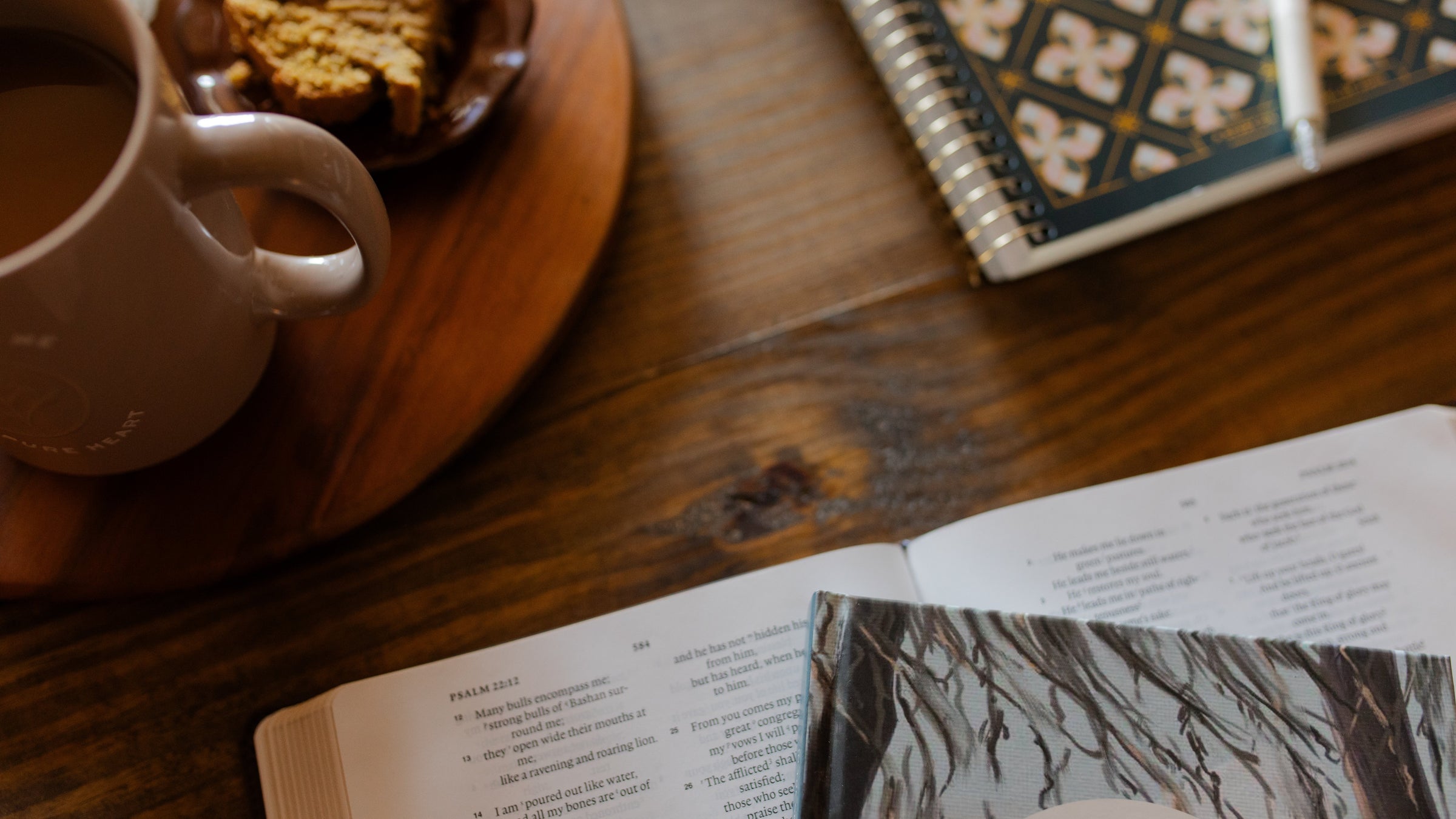 An open bible on a wooden table next to a wooden turn table with a coffee cup on it