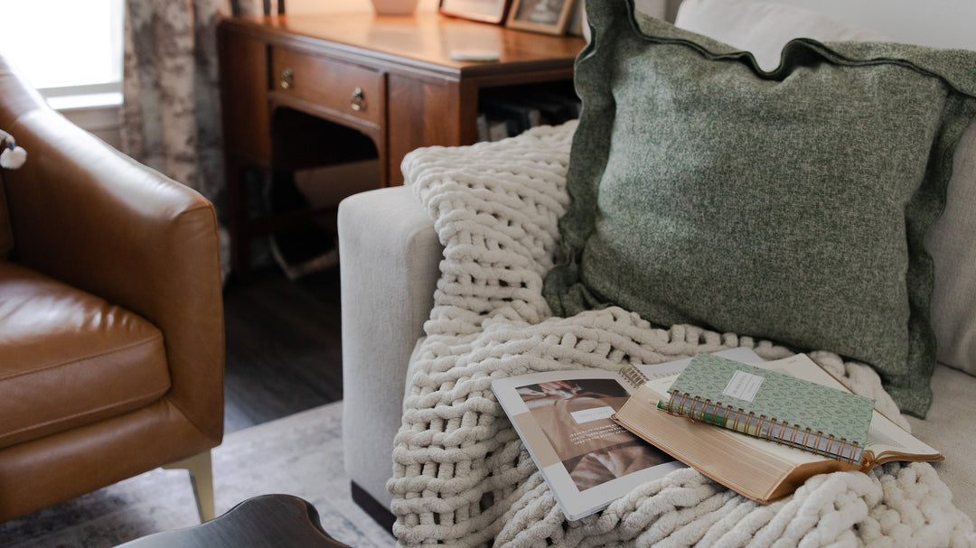 A white couch next to a leather couch, The white couch has a green pillow and a white knitted blanket on it along with various Daily Grace products