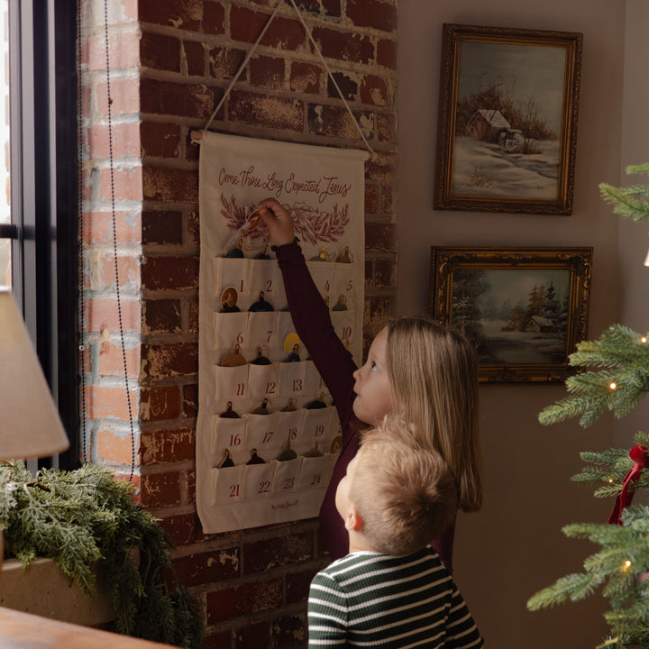 A child hangs a fabric Advent calendar on a brick wall in a cozy indoor setting, with another child standing beside them. The calendar has multiple small pockets containing small decorative items and numbered compartments.