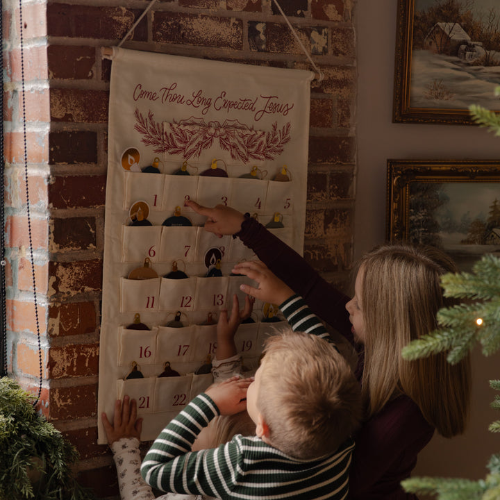 A woman and child hanging a fabric Advent calendar on a brick wall, with the woman pointing to one of the numbered pockets. The calendar has multiple small ornaments inside and the text 'Come Thou And Let Us Expect Thy Jesus' at the top.