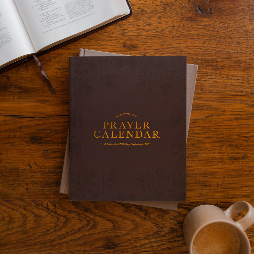 2026 Prayer Calendar on a closed journal on a wooden table next to an open bible and a coffee cup