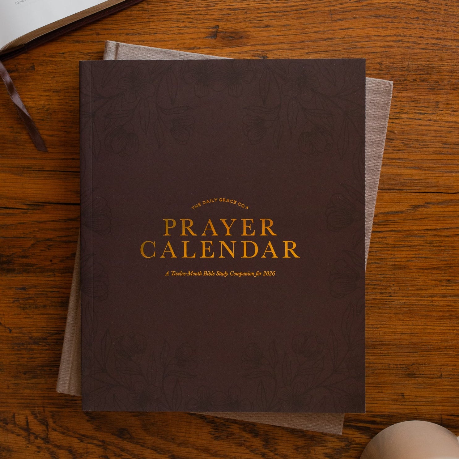 2026 Prayer Calendar on a closed journal on a wooden table next to an open bible and a coffee cup