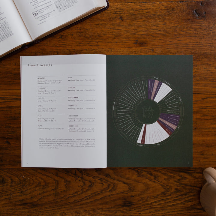 2026 Prayer Calendar opened to the Church seasons page on a wooden table next to an open bible and a coffee cup