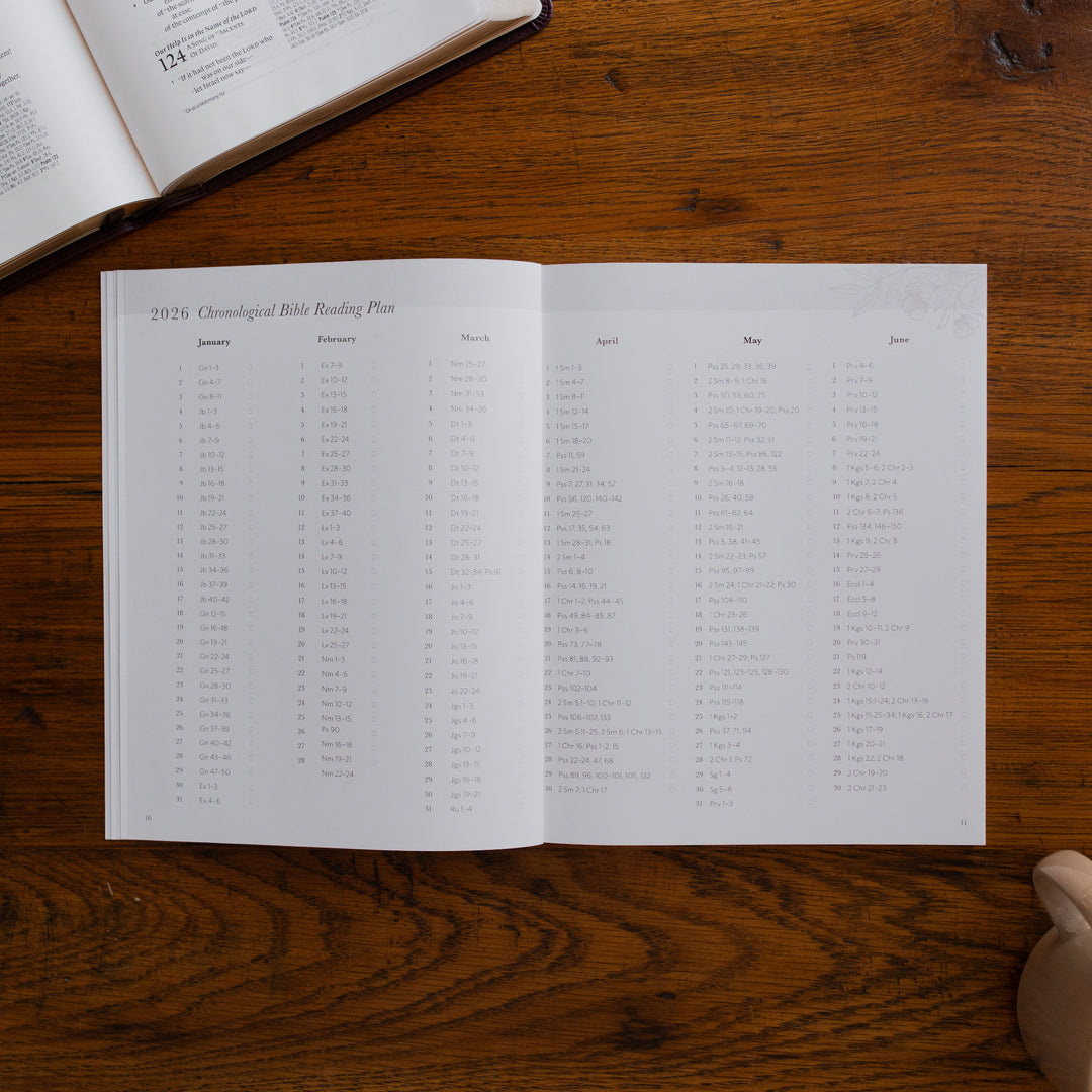 2026 Prayer Calendar opened to the chronological bible reading plan page on a wooden table next to an open bible