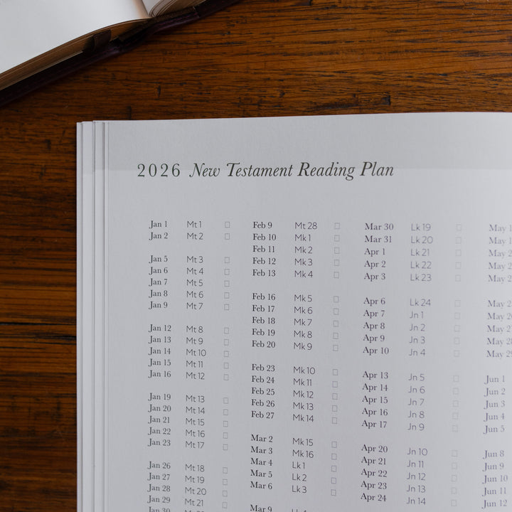 Up close of the 2026 Prayer Calendar opened to the new testament reading plan page on a wooden table next to an open bible