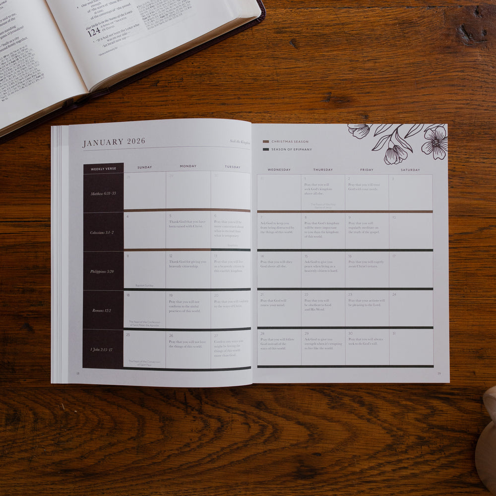 2026 Prayer Calendar opened to the January reading plan page on a wooden table next to an open bible