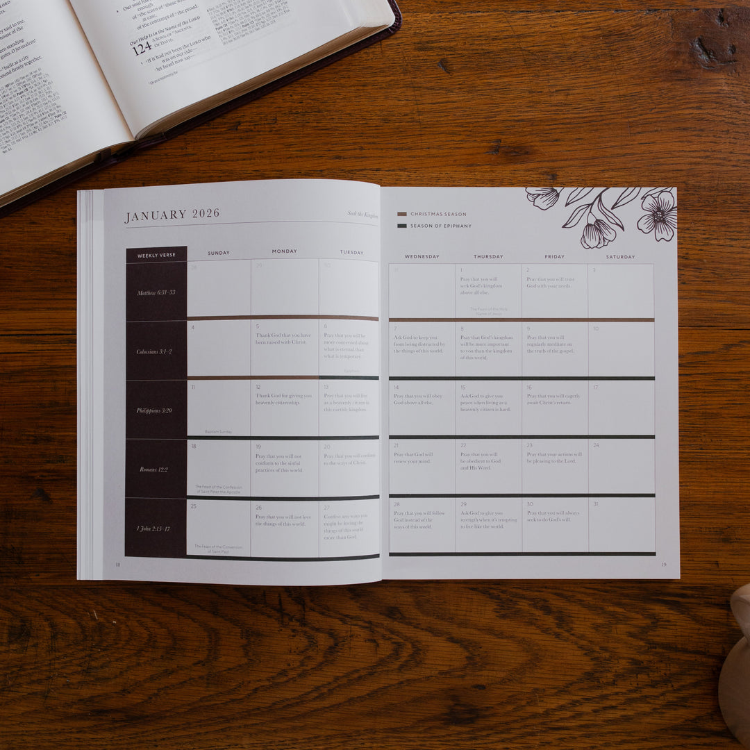 2026 Prayer Calendar opened to the January reading plan page on a wooden table next to an open bible