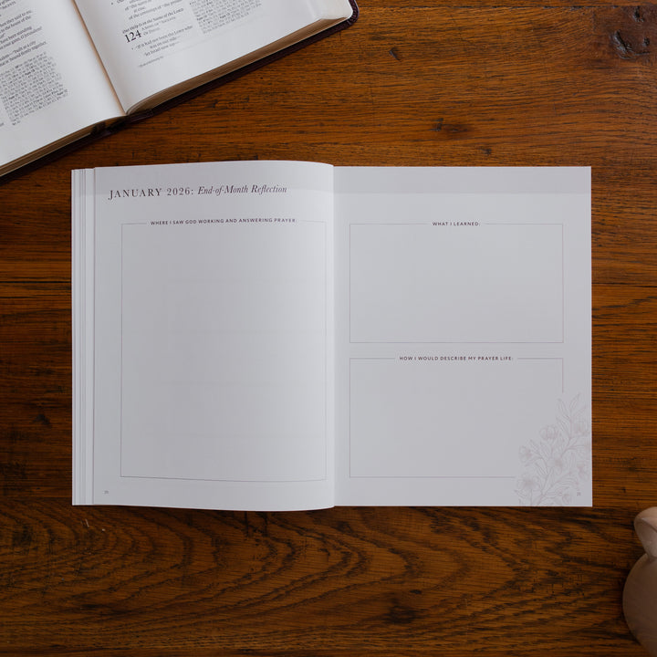 2026 Prayer Calendar opened to the end of month reflection page on a wooden table next to an open bible