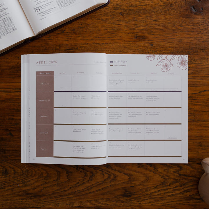 2026 Prayer Calendar opened to the April reading plan page on a wooden table next to an open bible