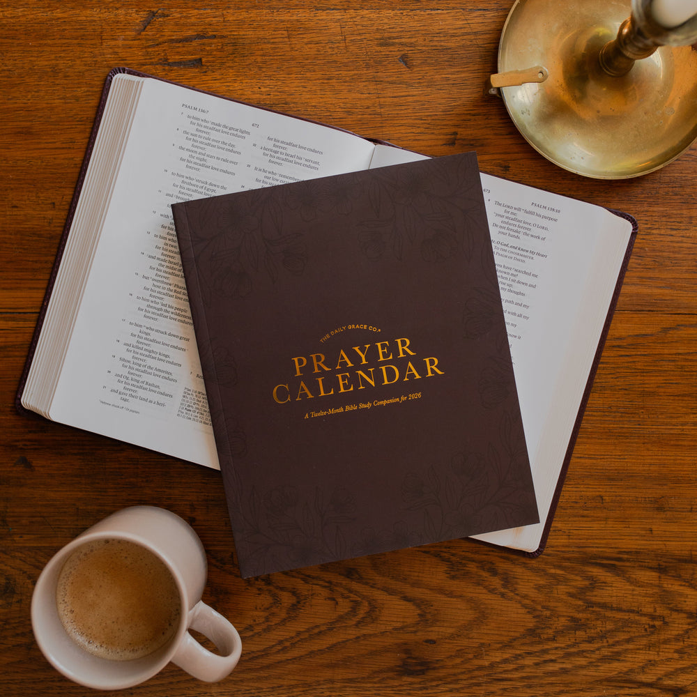 2026 Prayer Calendar on an open bible  on a wooden table next to a candle stick and a coffee cup