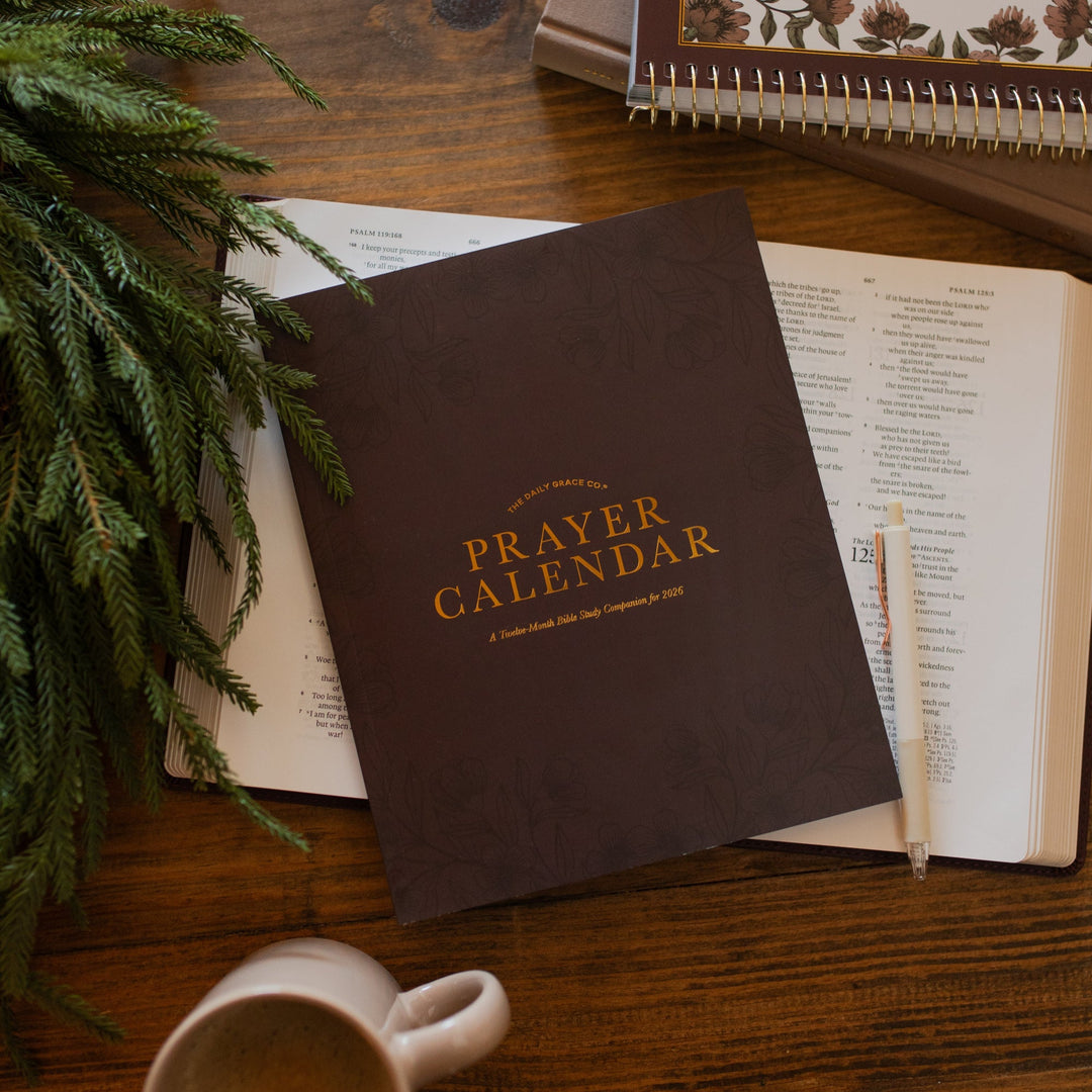 2026 Prayer Calendar on an open bible on a wooden table next to multiple closed books and a coffee cup