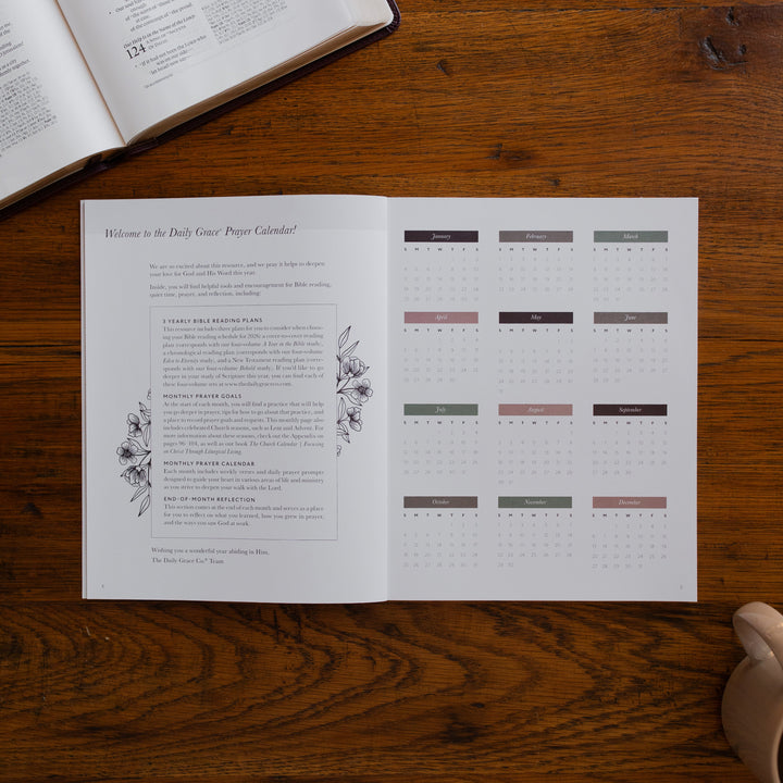 2026 Prayer Calendar opened to the welcome page on a wooden table next to an open bible