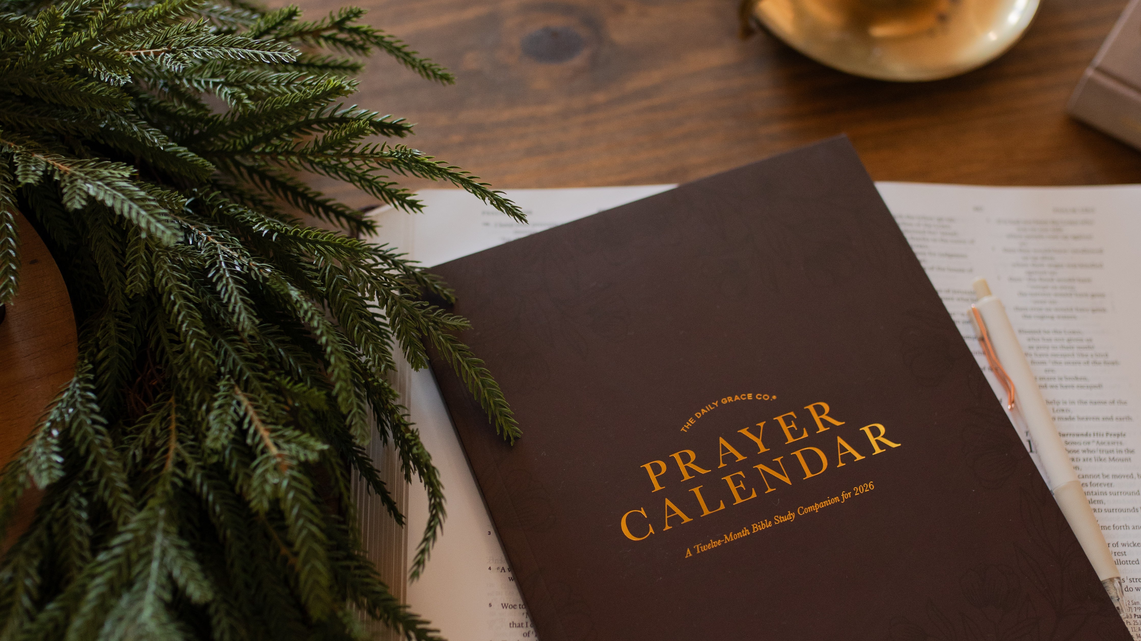 The 2026 Prayer Calendar on an open bible on a wooden table near some greenery