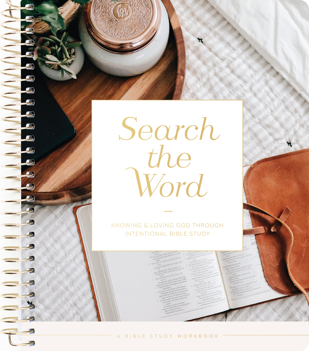 A spiral-bound Bible study workbook titled 'Search the Word' placed on a neutral fabric surface with an open book, a leather journal, and small decorative objects around it.