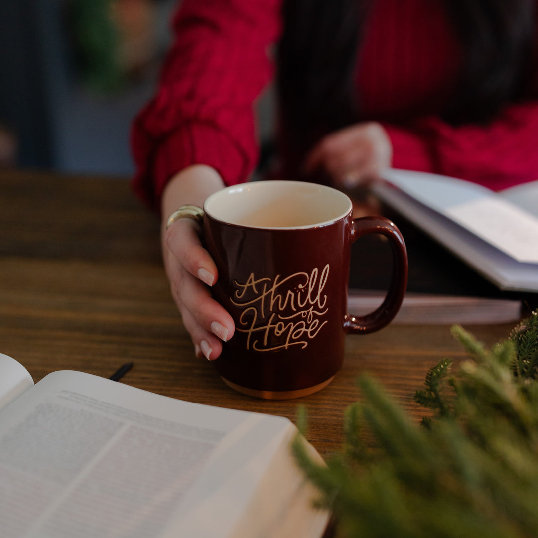 A woman holding onto the Thrill of Hope Mug resting on the wooden table next to an open bible