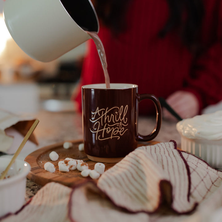 A woman pouring water into the Thrill of Hope Mug that is on a wooden round table with marshmallows on the table
