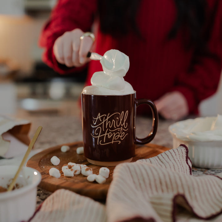 A woman pouring whipped cream into a Thrill of Hope Mug that is resting on a wooden round table with marshmallows next to the mug