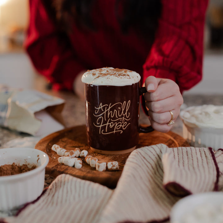 A woman holding the handle of the Thrill of Hope mug that is topped with whipped cream and cinnamon that is resting on a wooden round table next to some marshmallows